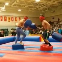 2 shirtless guys with safety gear hitting each other with foamed sticks