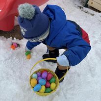 little kid holding a basket of colorful easter eggs picking up another egg