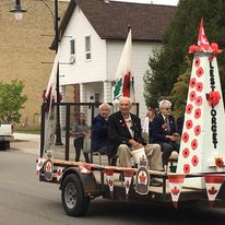 3 older men riding a cart with a white pyramid in front of it with flowers